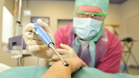 Ein Arzt in Schutzkleidung und Mundnasenschutz hält ein Instrument zur professionellen Wundreinigung in die Kamera und beugt sich dabei über einen liegenden Patienten, dessen Arm allein sichtbar ist.