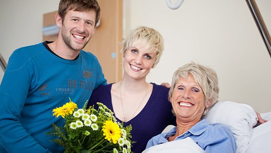Patienten & Besucher: Eine im Bett liegende Patientin wird von einer jungen Frau und einem Jungen Mann mit einem Blumenstrauß im Patientenzimmer besucht.